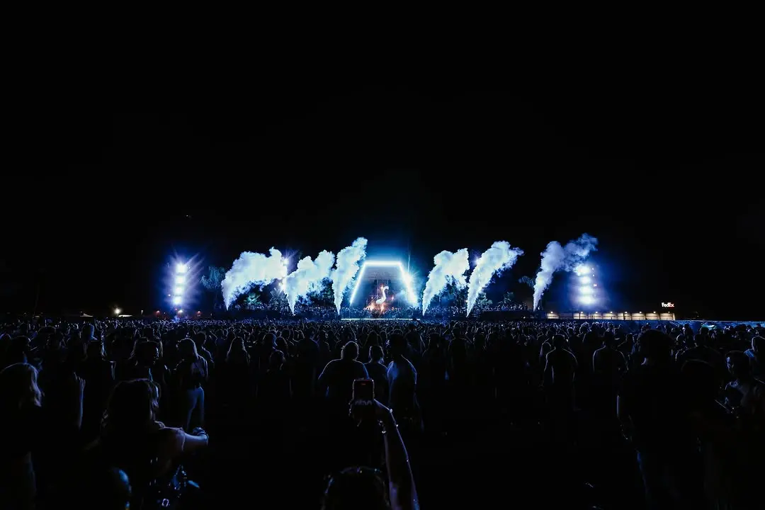 Vue large d'une scène de festival de nuit, jets de fumée bleue de part et d'autre d'une arche centrale, foule au premier plan.