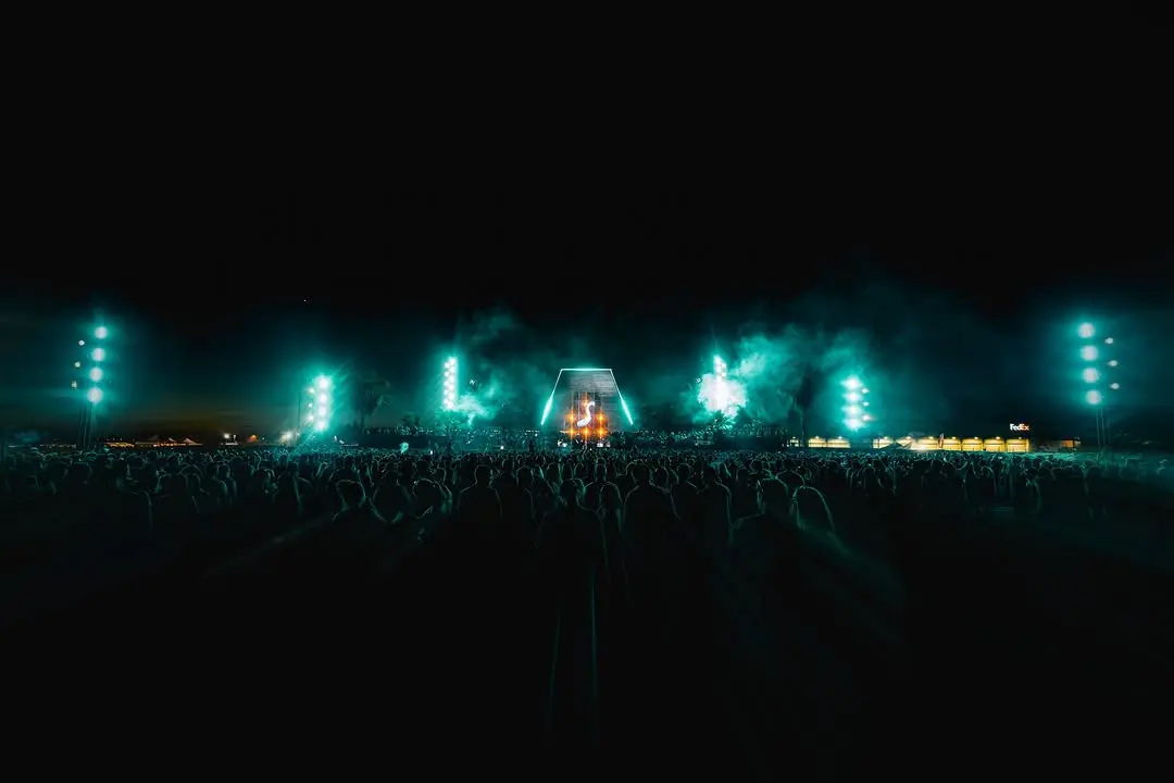 Scène de festival de nuit éclairée en vert émeraude, arche centrale et faisceaux verticaux, foule au premier plan.