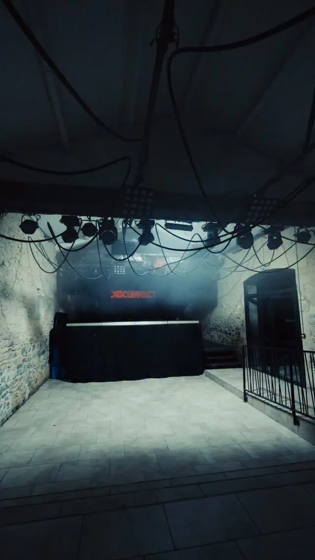 Intérieur d'un club en pierre, régie DJ sous une structure de lyres motorisées, enseigne néon rouge « DISCONNECT » dans la brume scénique.